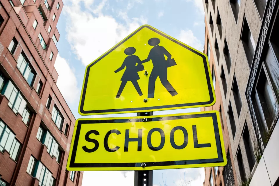 Yellow school crossing sign with two figures and brick buildings in background.